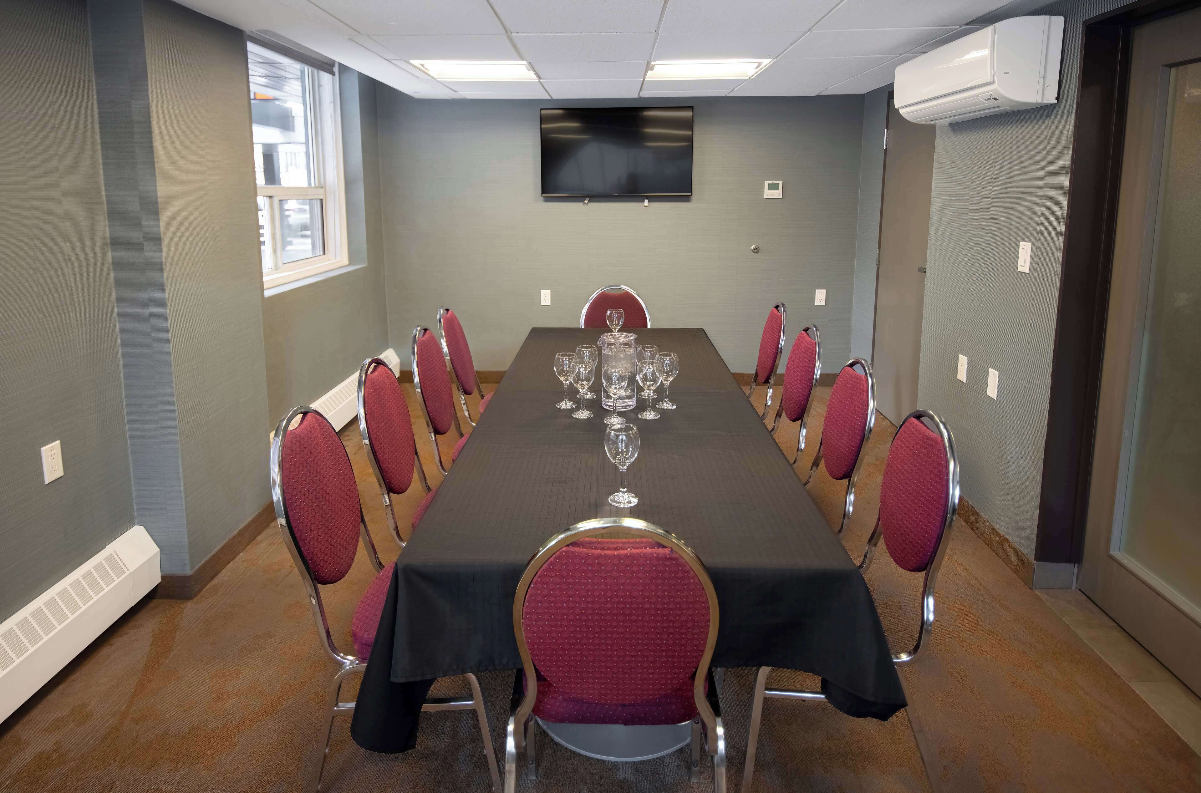 A conference room with a long table, red chairs, a TV on the wall, and an air conditioning unit.