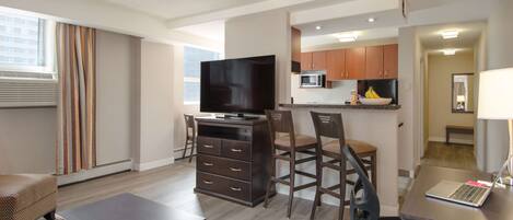 Una habitación de hotel moderna con televisión de pantalla plana, zona de comedor con mesa y sillas, kitchenette con microondas y cocina, y un amplio ventanal con cortinas.