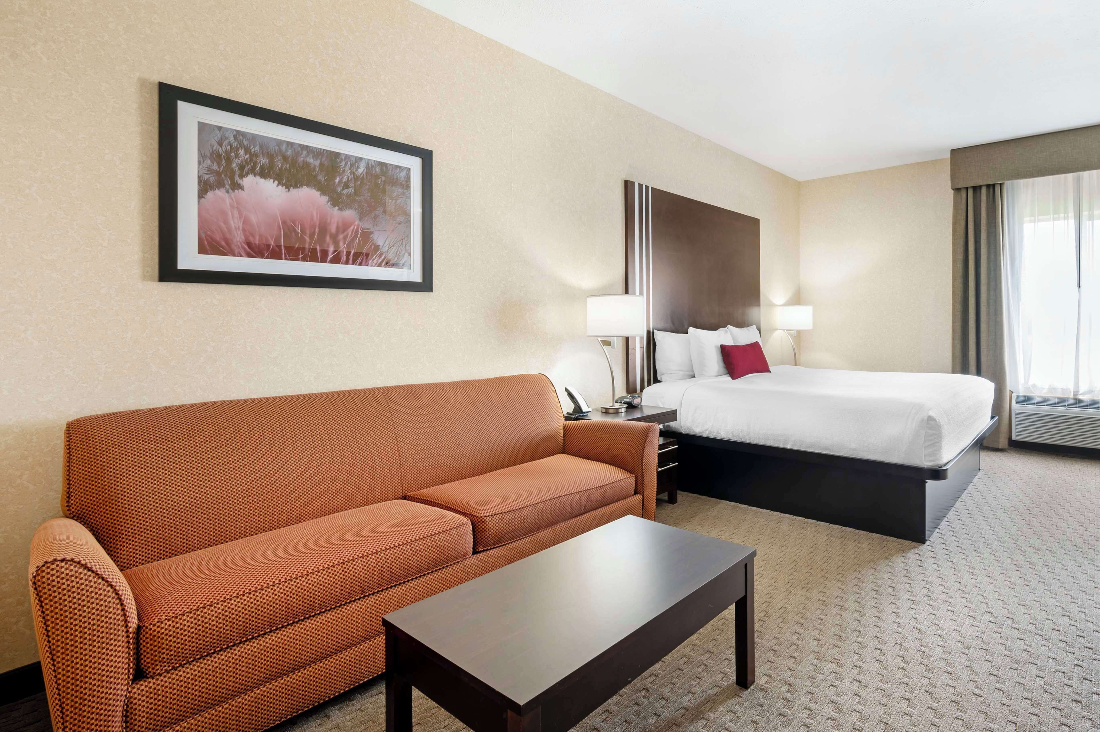A hotel room with a brown sofa, a bed with white linens, a bedside table, a lamp, and a framed picture on the wall.