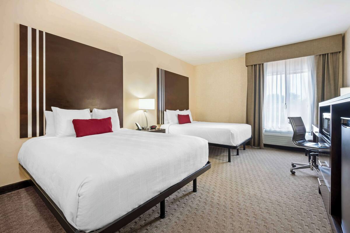 A hotel room with two beds, a desk with a chair, a TV, and a large window with curtains.