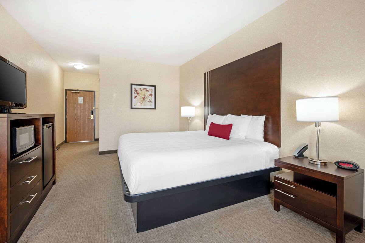 A hotel room with a large bed, a bedside table with a lamp, a TV, and a minibar.