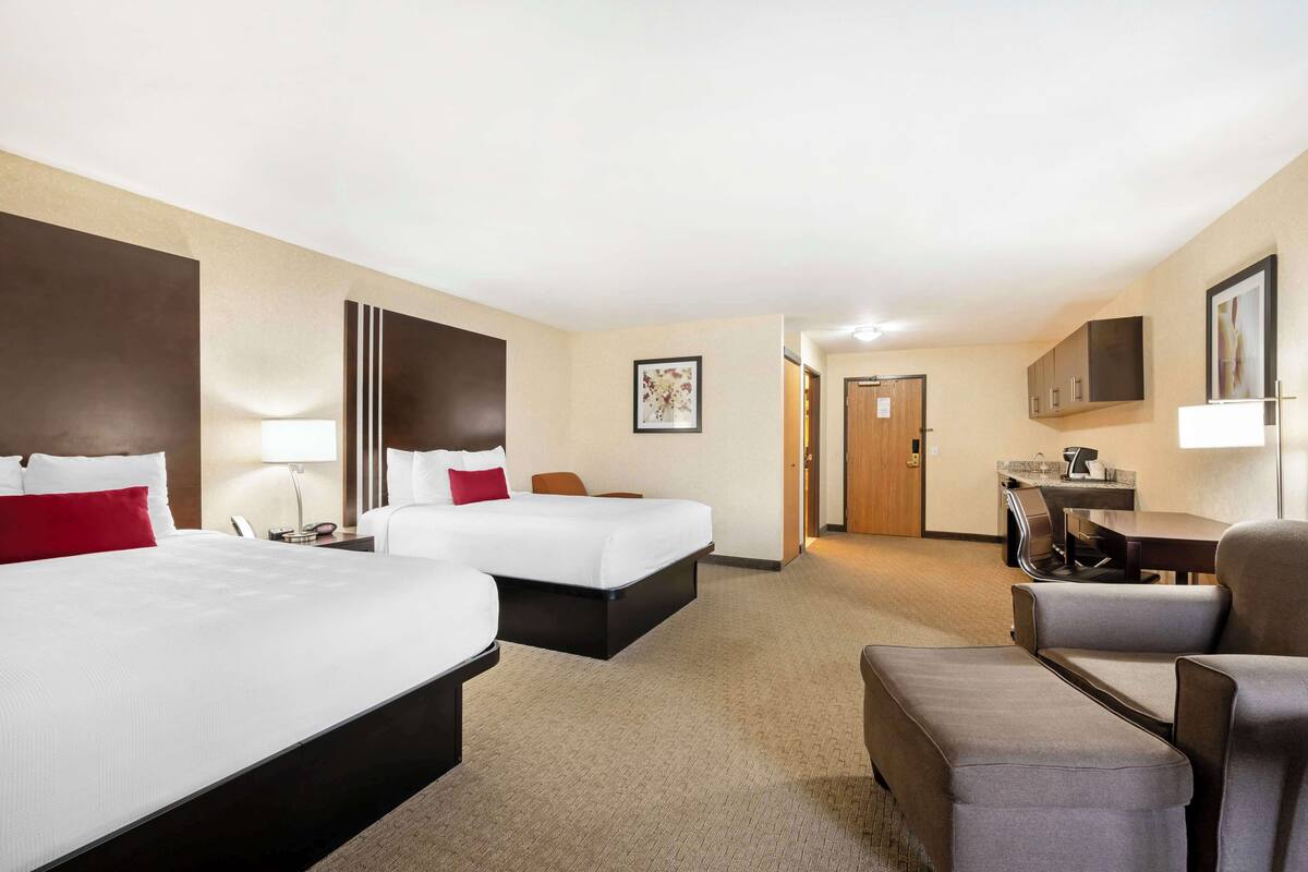 A hotel room with two beds, a desk, a chair, and a small kitchenette.