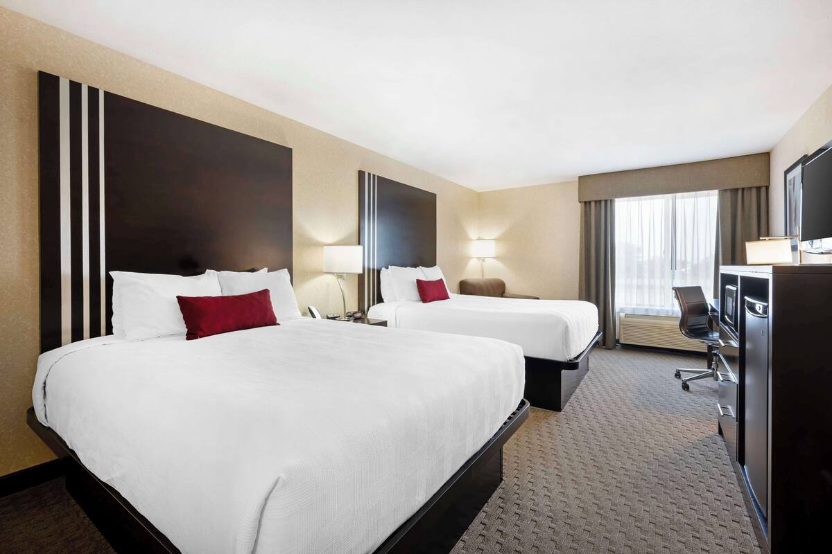A hotel room with two beds, a desk, a TV, and a large window with curtains.