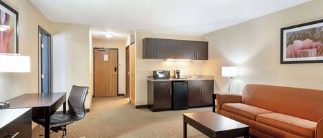 A hotel room with a desk, office chair, sofa, coffee table, kitchenette, and a framed picture on the wall.
