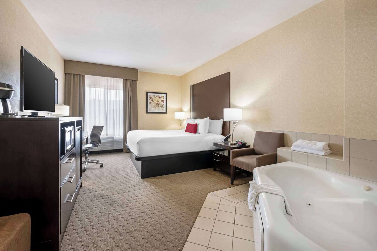 A hotel room with a bed, a desk with a computer, a TV, a chair, a lamp, a bathtub, and a window with curtains.
