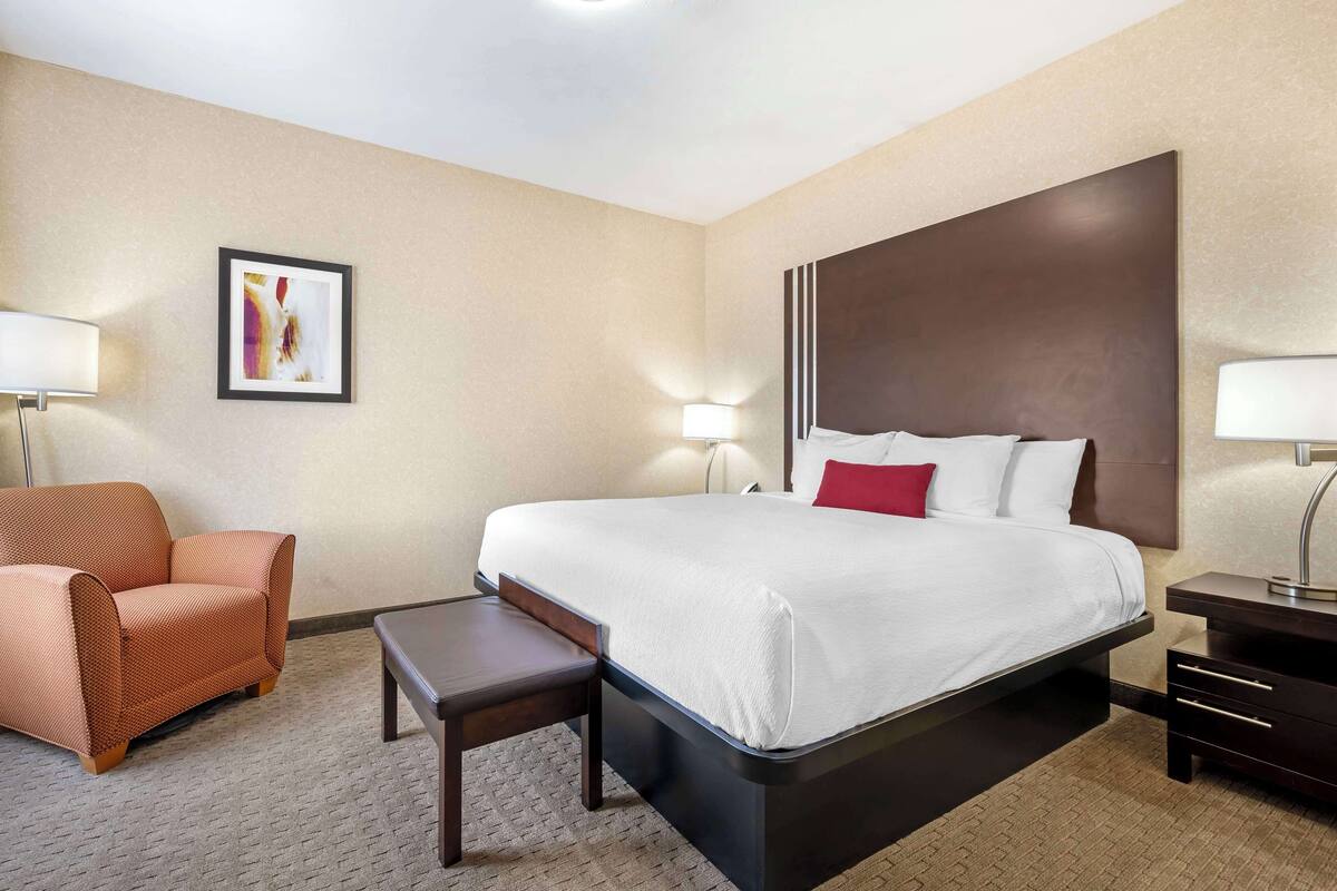 A hotel room with a large bed, a chair, a small table, and a nightstand with a lamp.