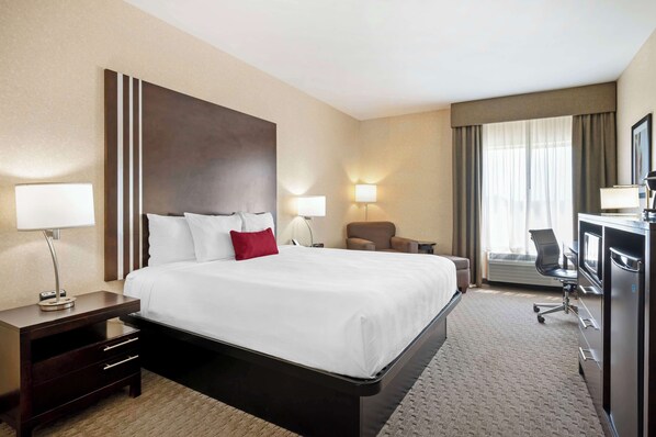 A hotel room with a large bed, two bedside tables with lamps, a desk with a chair, and a large window with curtains.