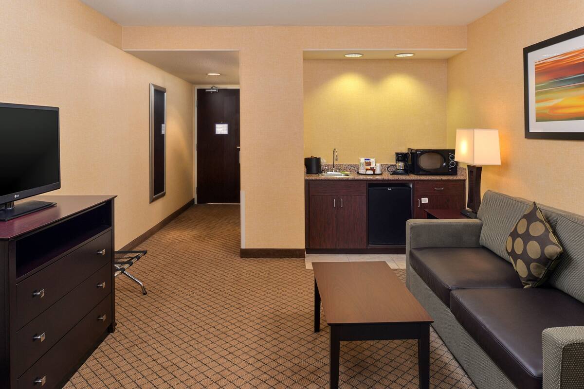 A hotel room with a television on a dresser, a sofa, a small table, a kitchenette with a microwave, and a framed picture on the wall.