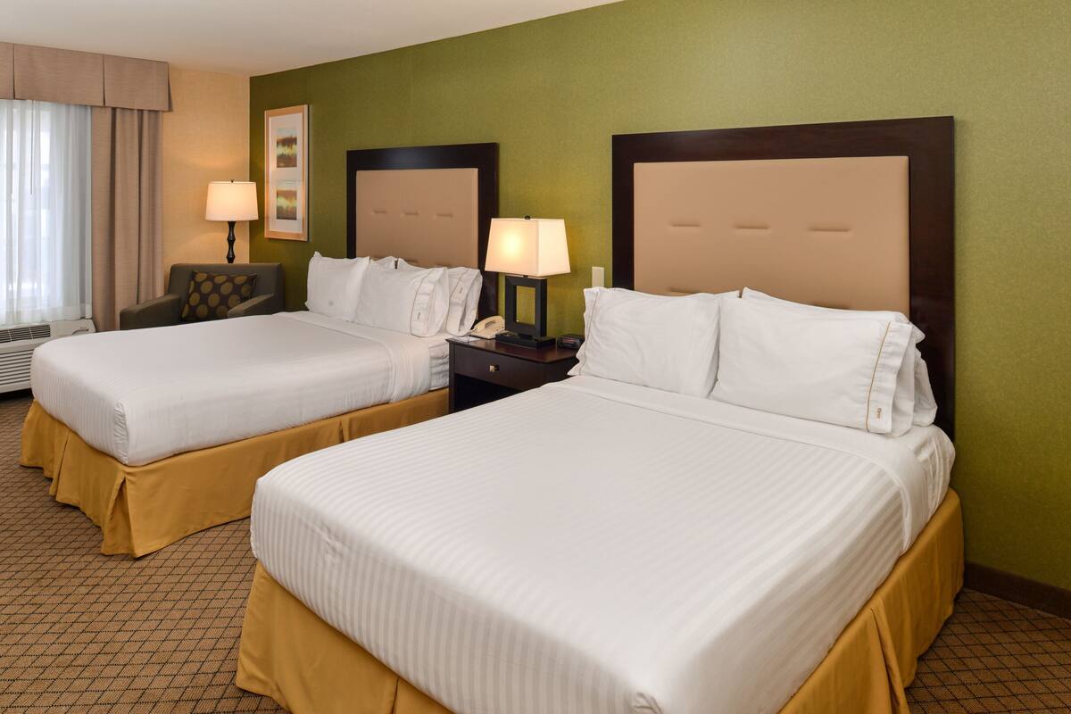 A hotel room with two beds, a nightstand with a lamp, a chair, and a window with curtains.