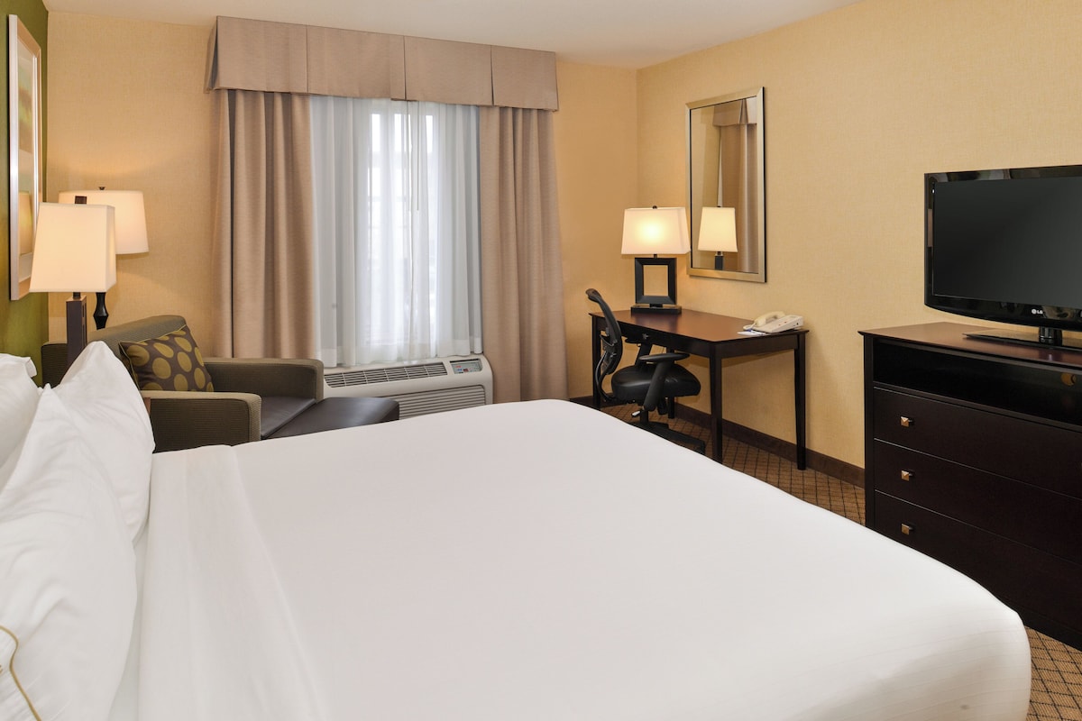 Una habitación de hotel con una cama grande, un escritorio con silla, televisor y ventana con cortinas.