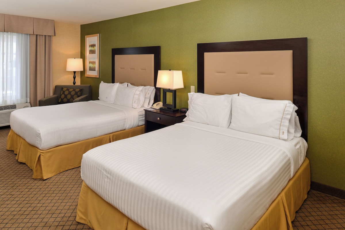 Habitación de hotel con dos camas, una mesita de noche con lámpara, una silla y un ventanal con cortinas.