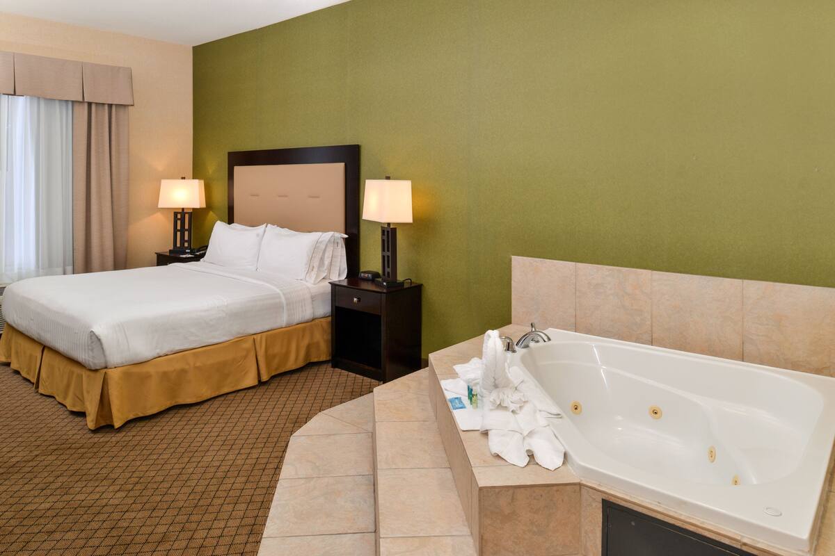 A hotel room with a bed, two bedside tables with lamps, a large bathtub, and a green wall.