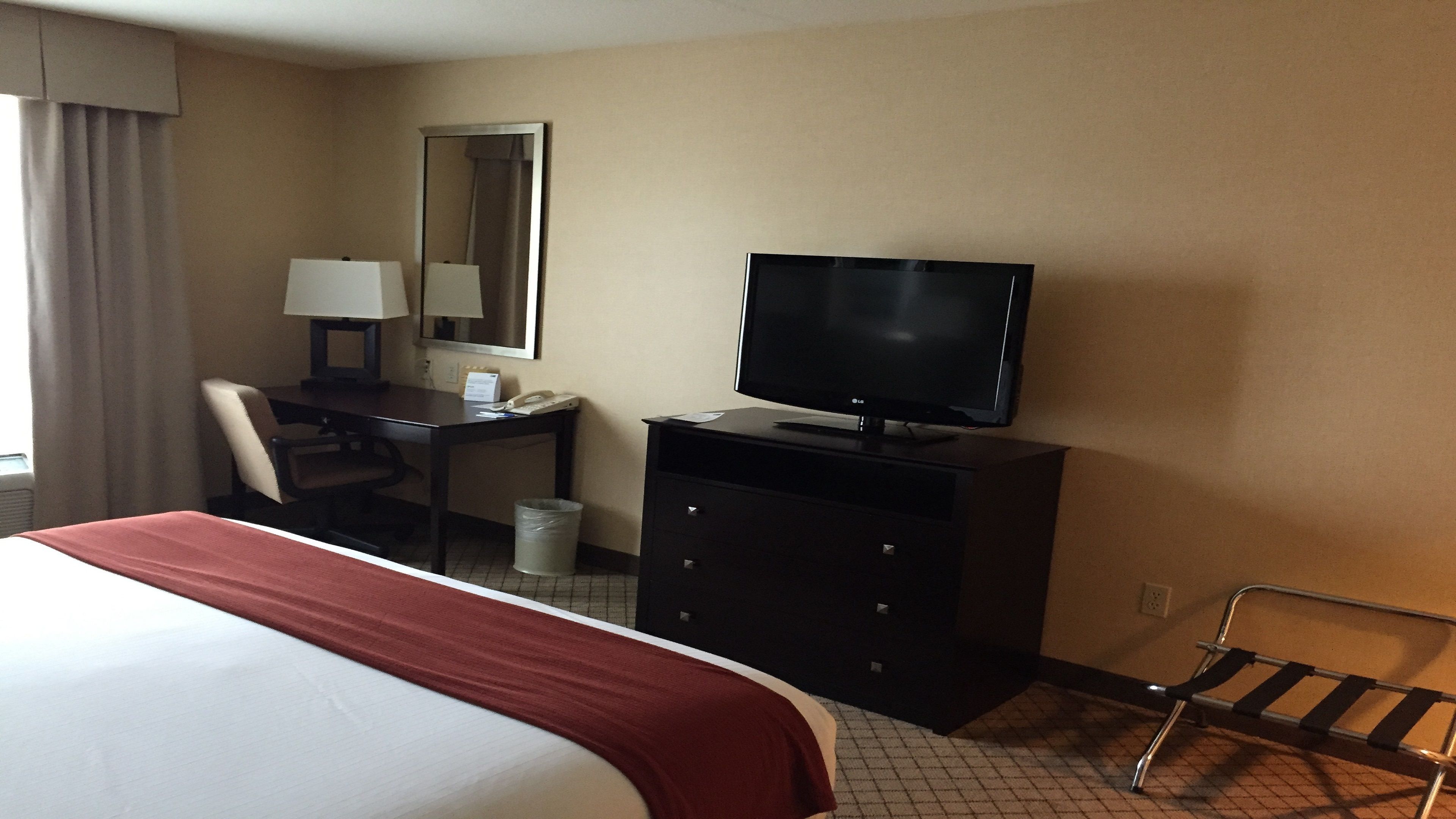 A hotel room with a bed, a desk with a chair, a television, a mirror, and a trash bin.