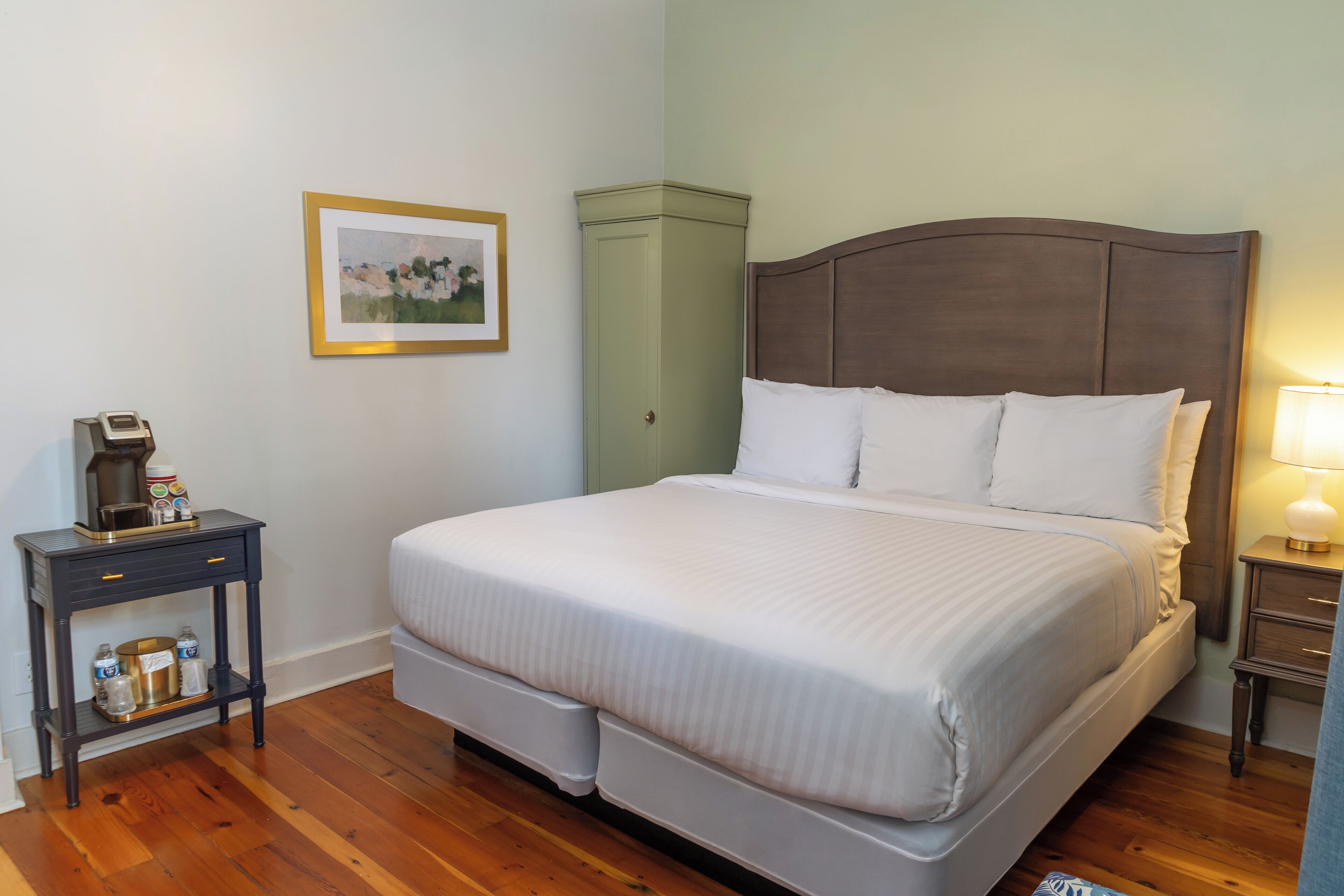 Una cama bien hecha con sábanas blancas, una mesita de noche con una lámpara y un cuadro en la pared.