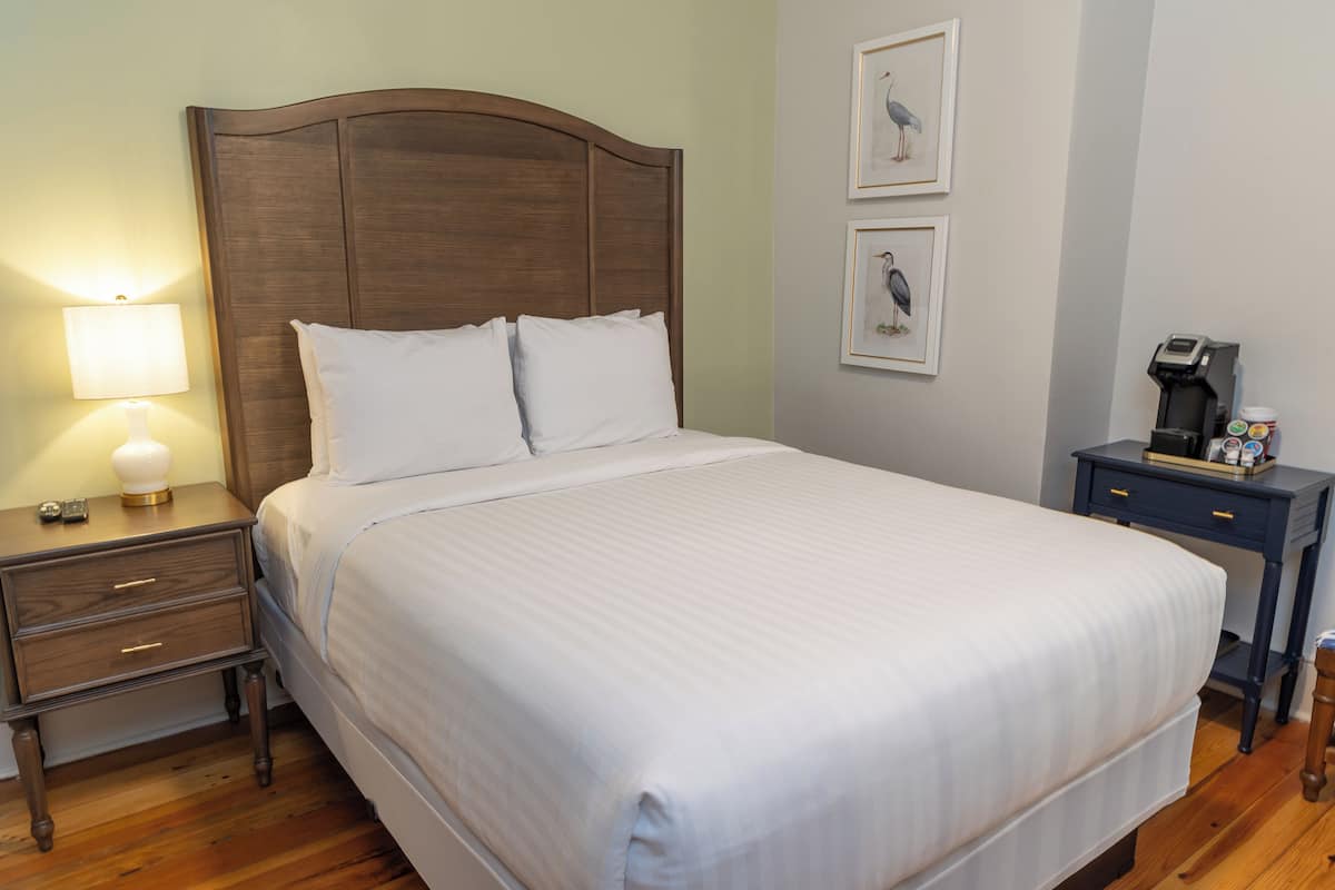 Una cama bien hecha con sábanas blancas, cabecera de madera, mesita de noche con lámpara y cafetera sobre una pequeña mesa al lado de la cama.