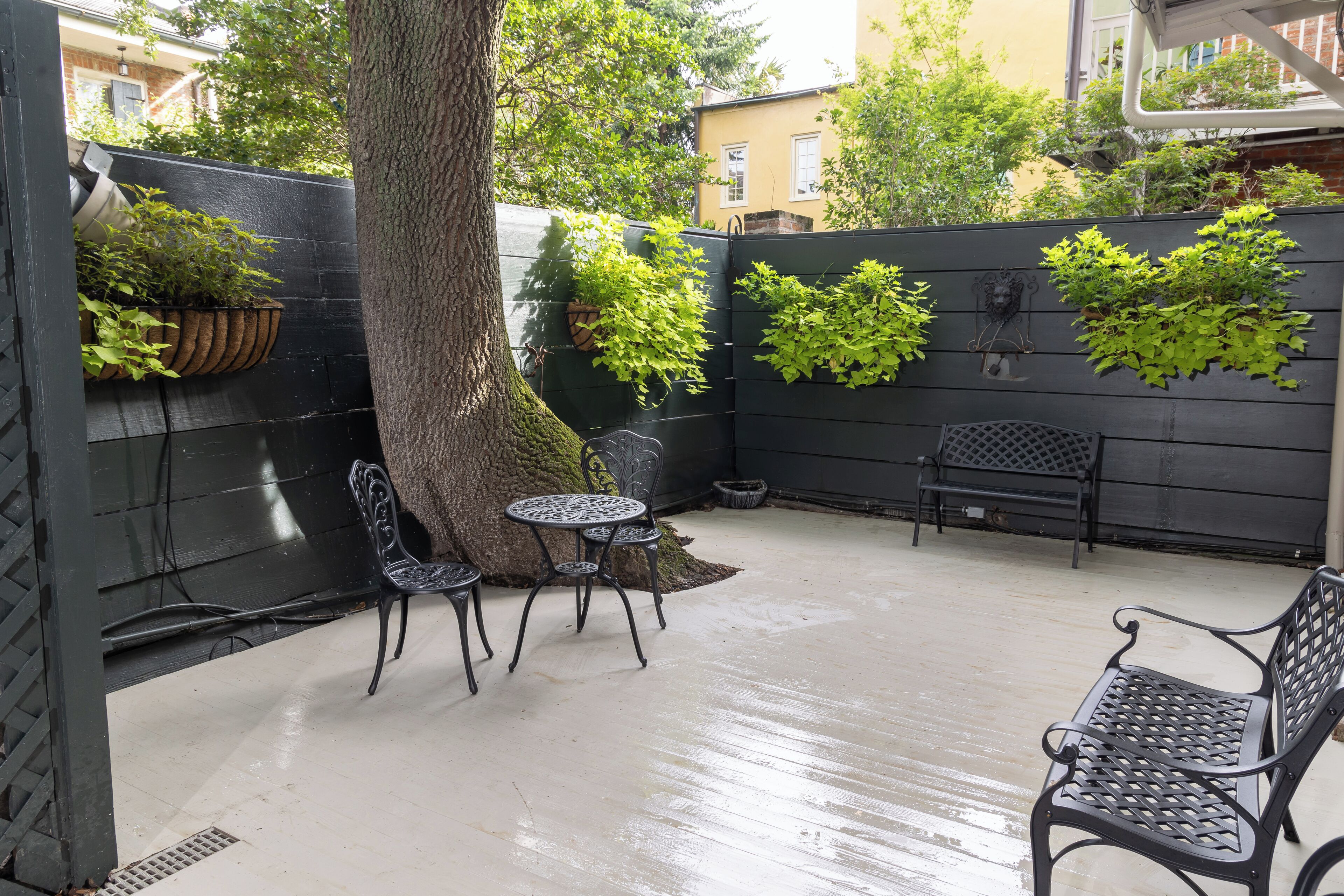Een patio met een zwart metalen bankje, een ronde metalen tafel en stoelen. Er staan potplanten tegen de muur en staat een boomstam.