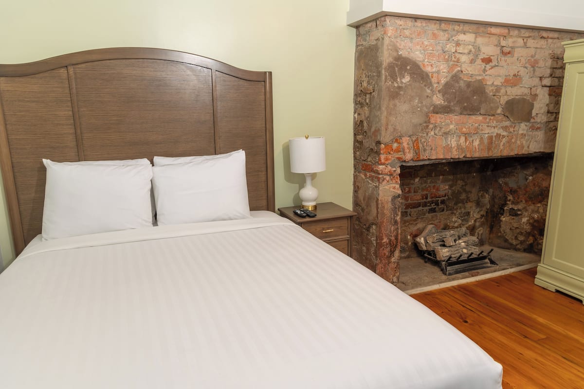 Una cama bien hecha con sábanas blancas, un cabecero de madera, una mesita de noche con lámpara y una chimenea de ladrillo.