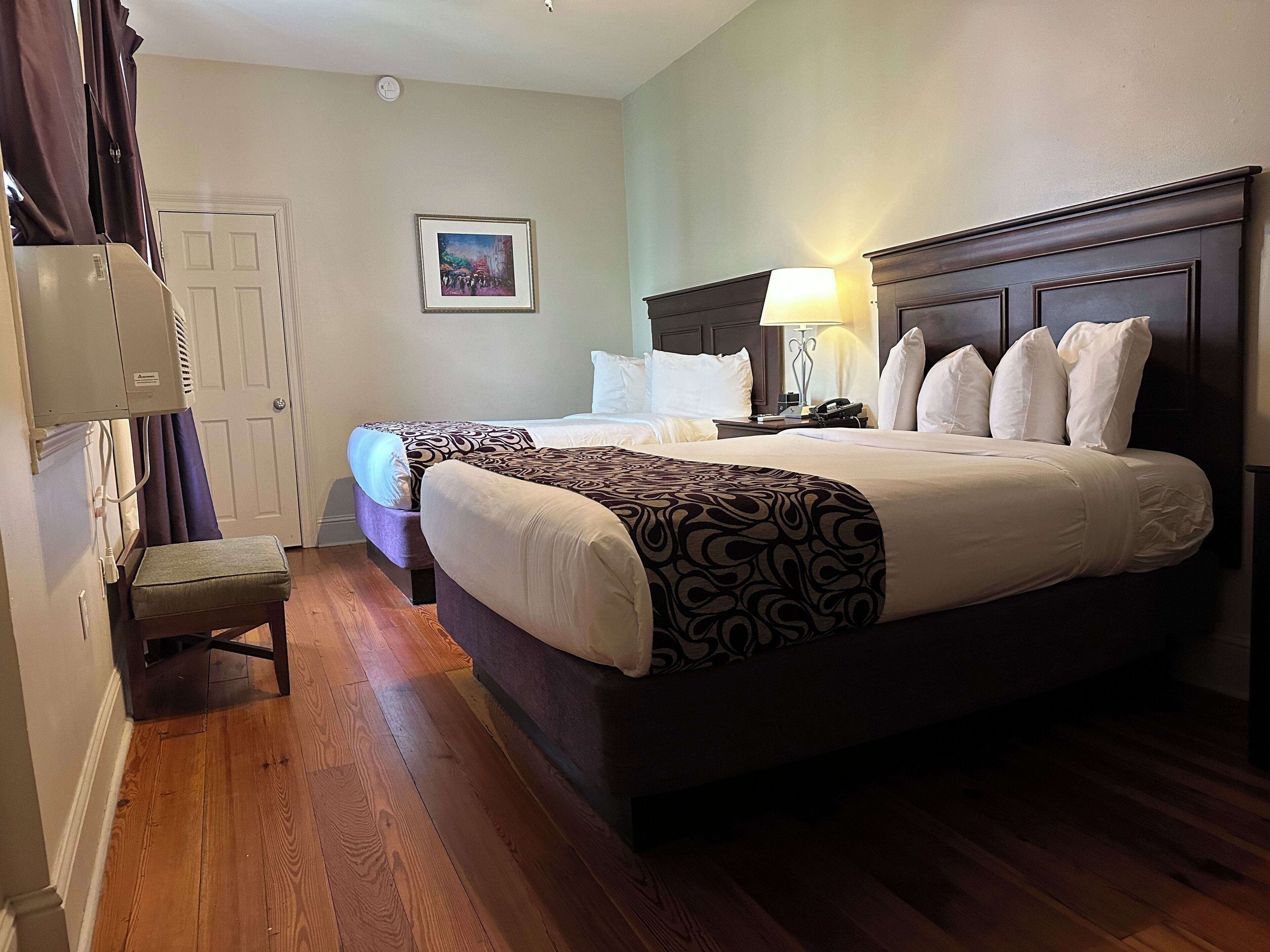A hotel room with a large bed, a smaller bed, a nightstand with a lamp, a chair, a door, and a wall-mounted air conditioning unit.