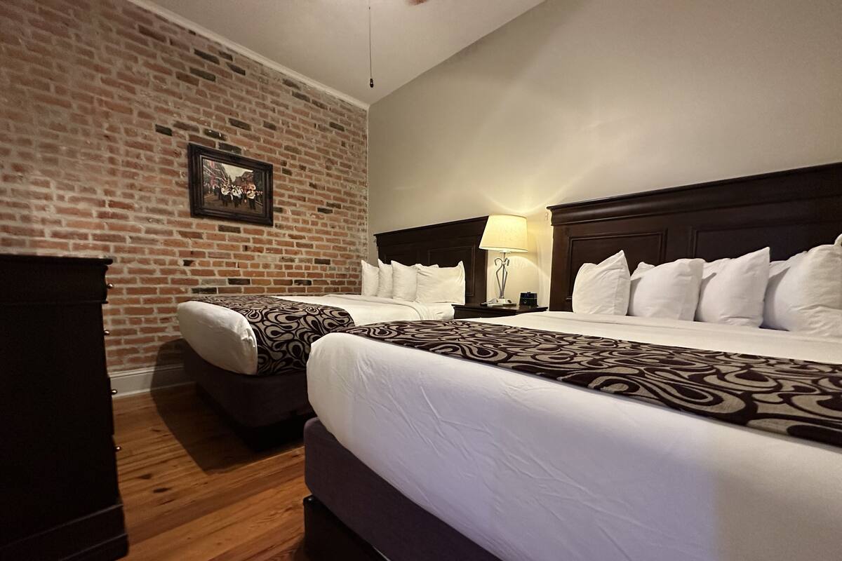 A hotel room with two beds, a brick wall, a wooden floor, and a nightstand with a lamp.