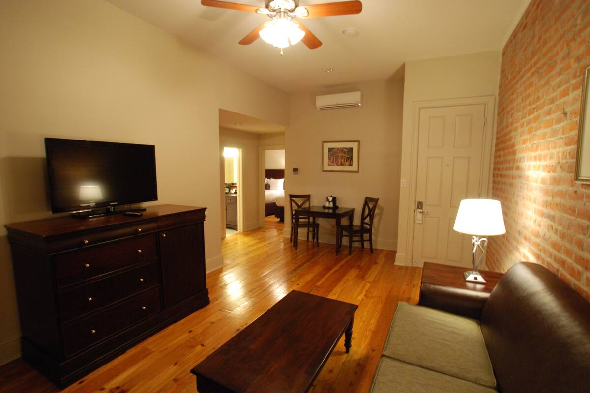 A living room with a wooden floor, a dark wooden cabinet, a flat-screen TV, a brown leather sofa, a wooden coffee table, a dining table with chairs, and a ceiling fan.