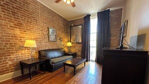 A living room with a brick wall, a leather sofa, a wooden coffee table, a TV on a cabinet, and two lamps. - Inn on St. Peter (New Orleans)