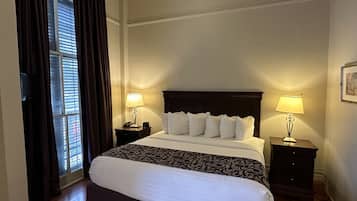 A hotel room with a large bed, two bedside tables with lamps, a window with curtains, and a wooden floor.