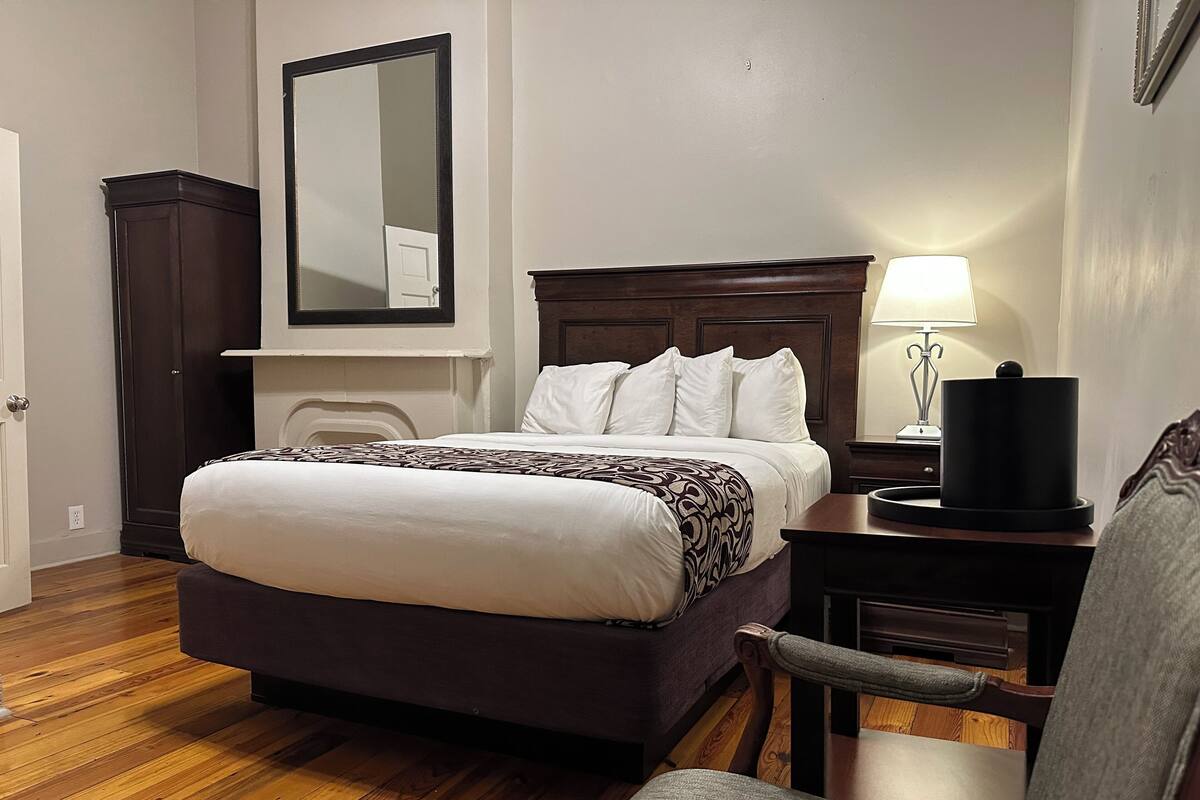 A hotel room with a large bed, a nightstand with a lamp, a chair, a wardrobe, and a mirror on the wall.