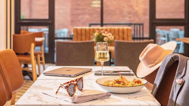 Une table à manger avec un ordinateur portable, un verre à vin, une assiette de nourriture et une paire de lunettes de soleil.