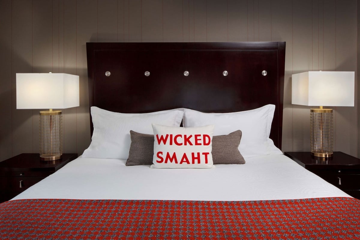 Une chambre d’hôtel avec un grand lit, deux tables de chevet avec des lampes, une tête de lit sombre et un oreiller orné du texte « WICKED SMART ».
