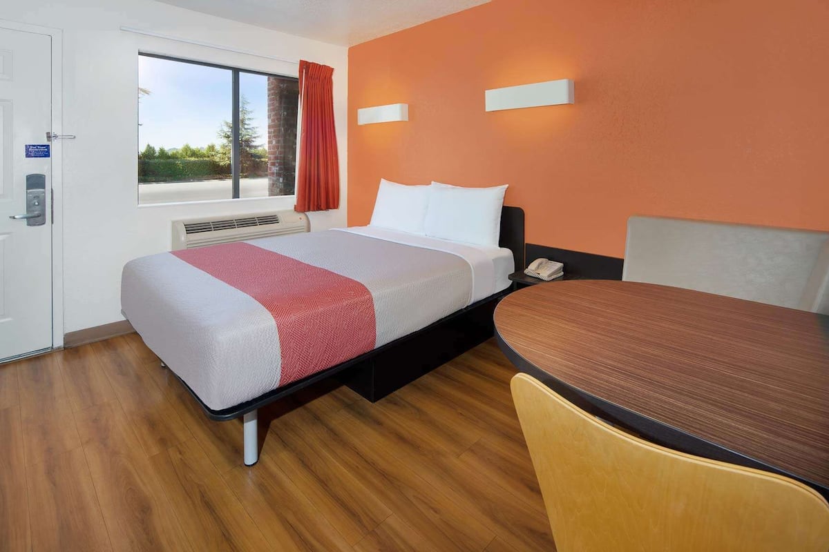 A hotel room with a bed, a desk, and a chair. There is a window with a view of trees and a building.
