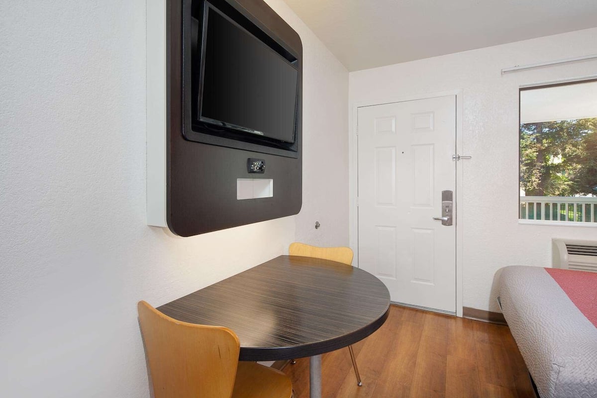 A room with a round table, two chairs, a mounted TV, a door, and a window with a view of trees.