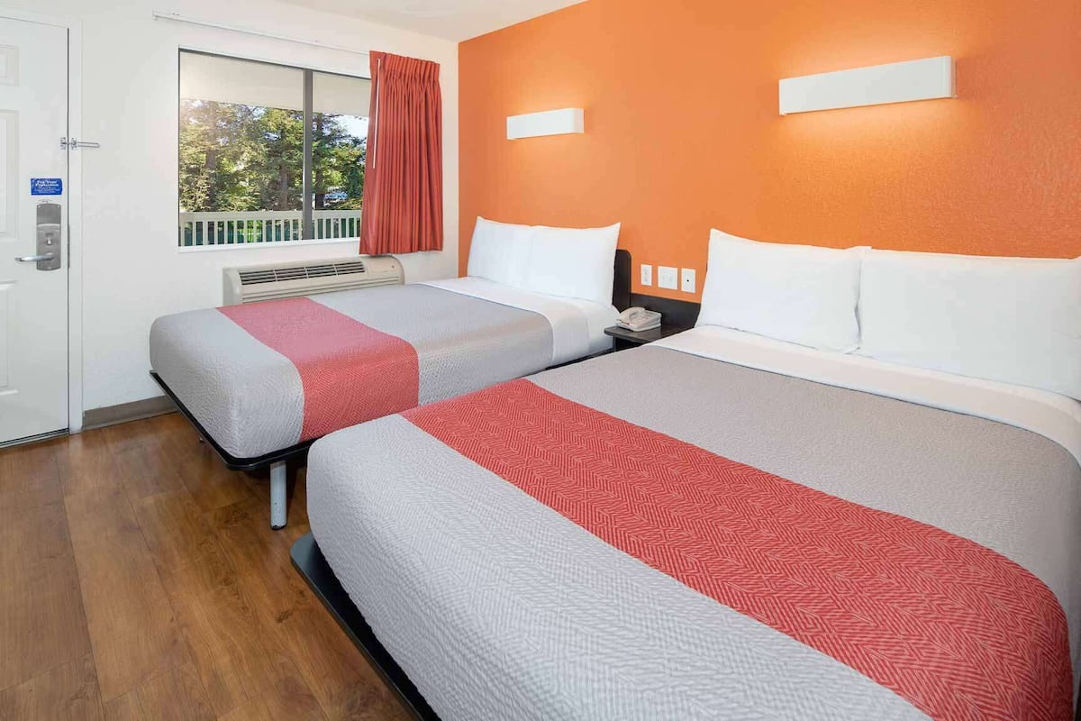 A hotel room with two beds, a window with curtains, a thermostat, and a phone on the nightstand.