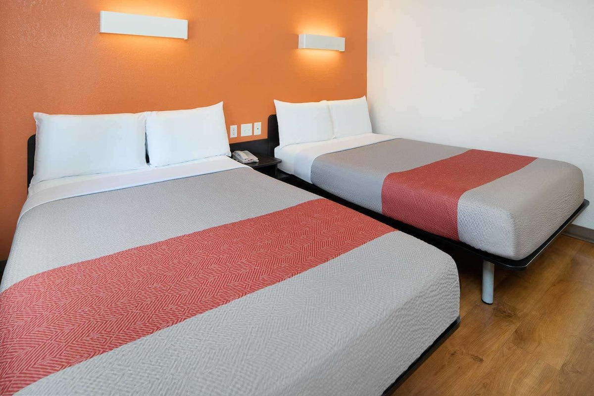A hotel room with two beds, each with white linens and a red and grey bedspread. There are wall-mounted lights above the beds and a nightstand with a phone.