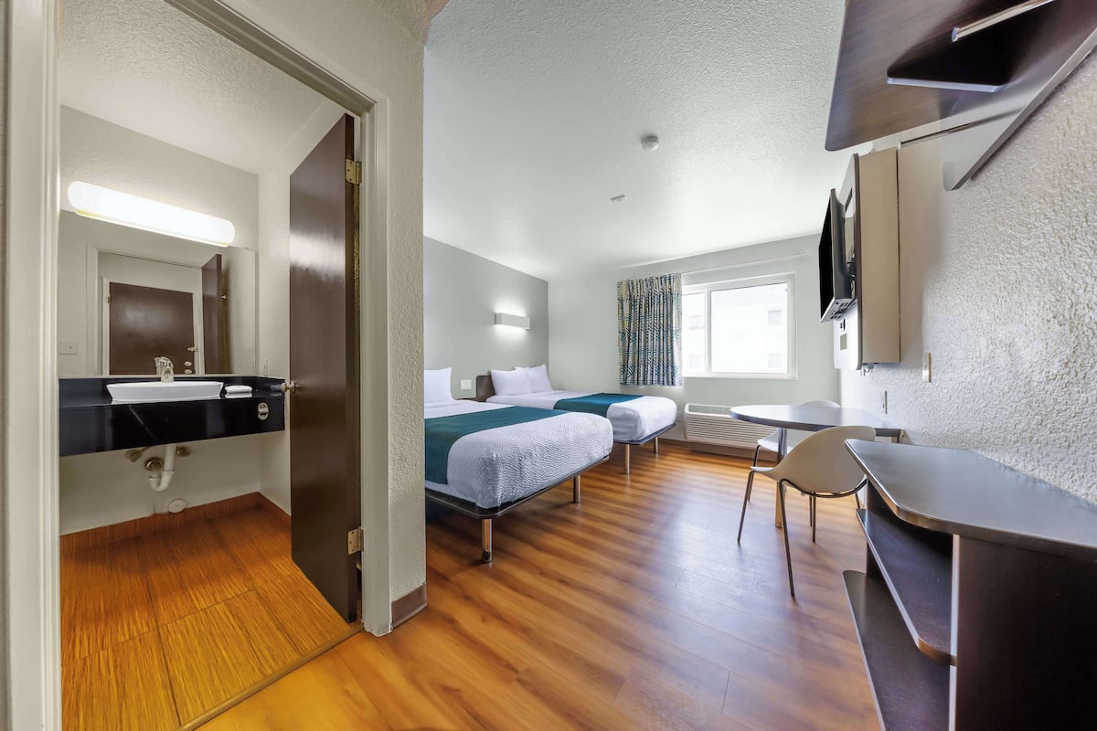 A hotel room with a wooden floor, a bed with a green blanket, a desk, a chair, a TV mounted on the wall, and a window with curtains.