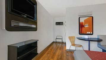 Quarto de hotel moderno com televisão de tela plana, uma pequena mesa com uma cadeira, uma cama com colcha estampada em vermelho e cinza, e um espelho na parede.