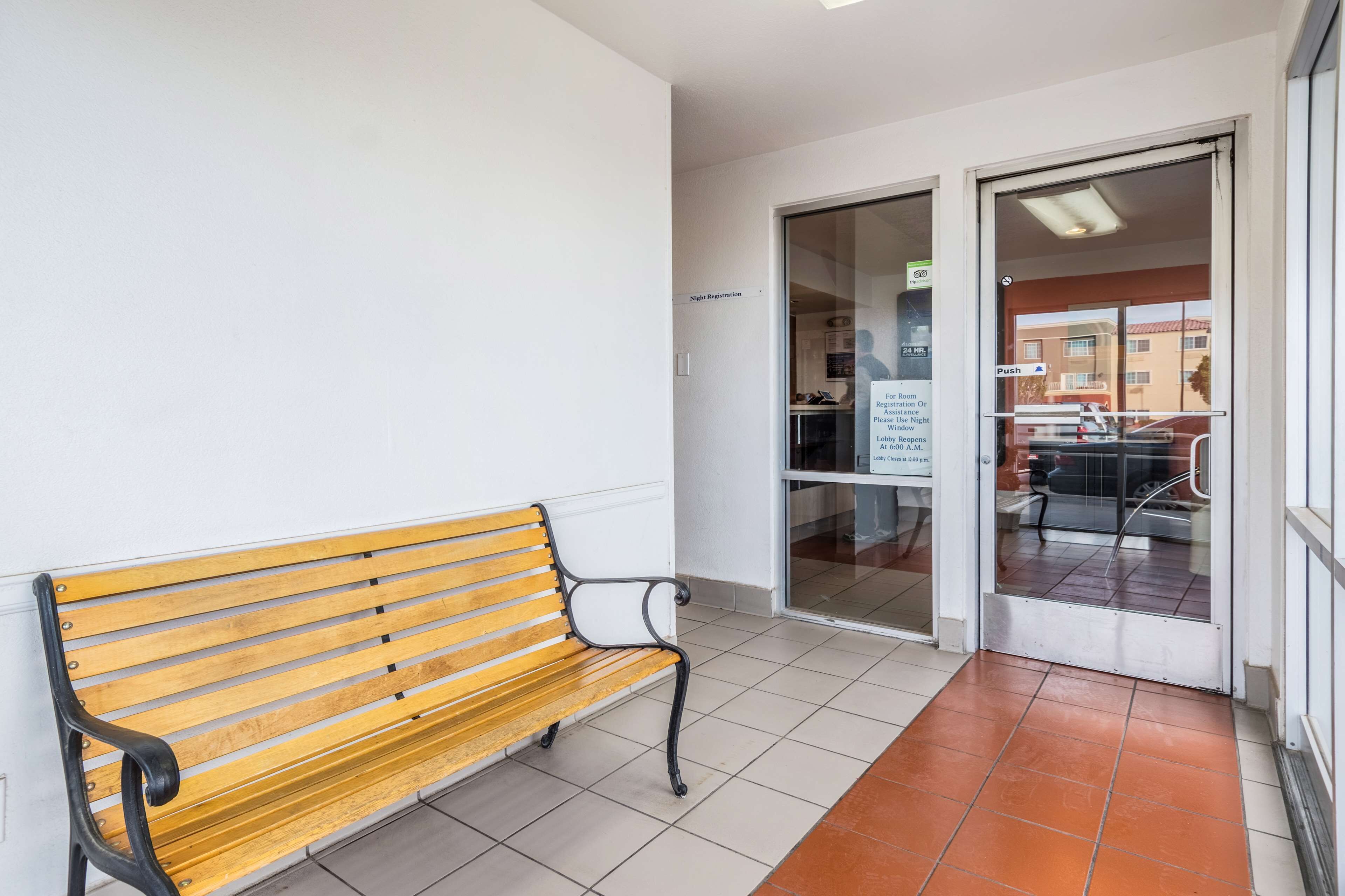 Een houten bank in een gang met een betegelde vloer, een glazen deur die uitkomt in een lobby en een bord met nachtregistratie.