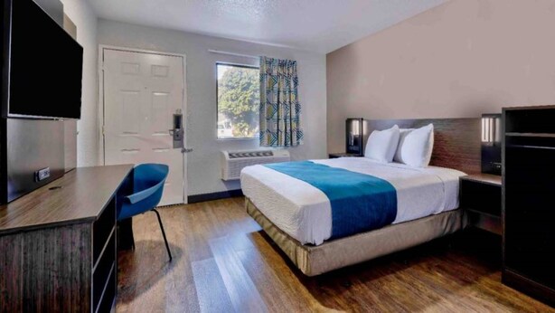 Habitación de hotel con cama, escritorio, silla, televisor y ventana con cortinas.