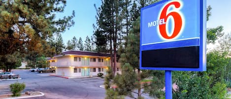Señal de motel con el número 6, un edificio de dos pisos con techo rojo y un estacionamiento con varios autos.