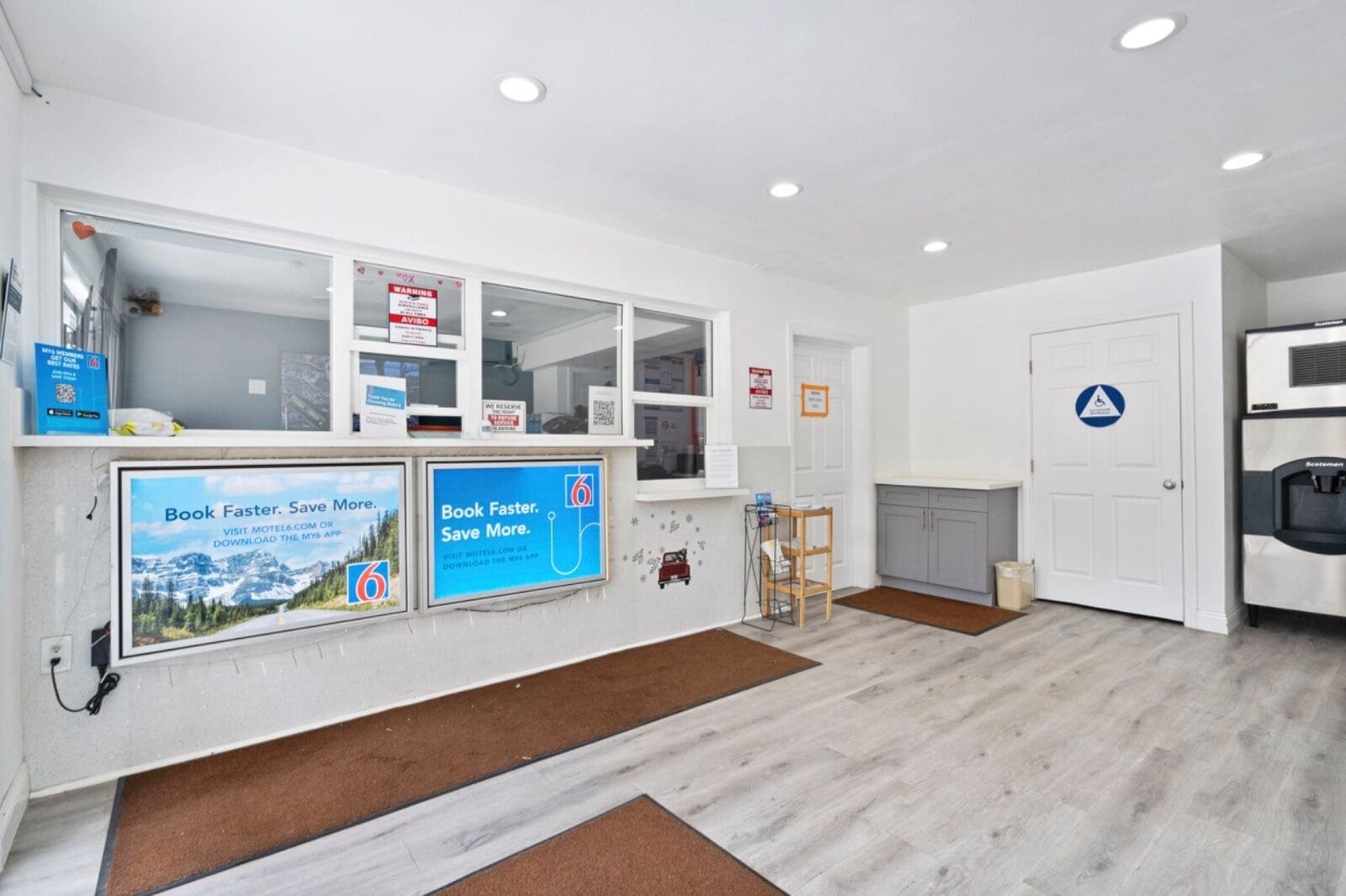 A clean, modern hotel lobby with a reception desk, informational posters, a water dispenser, and a wheelchair-accessible sign.