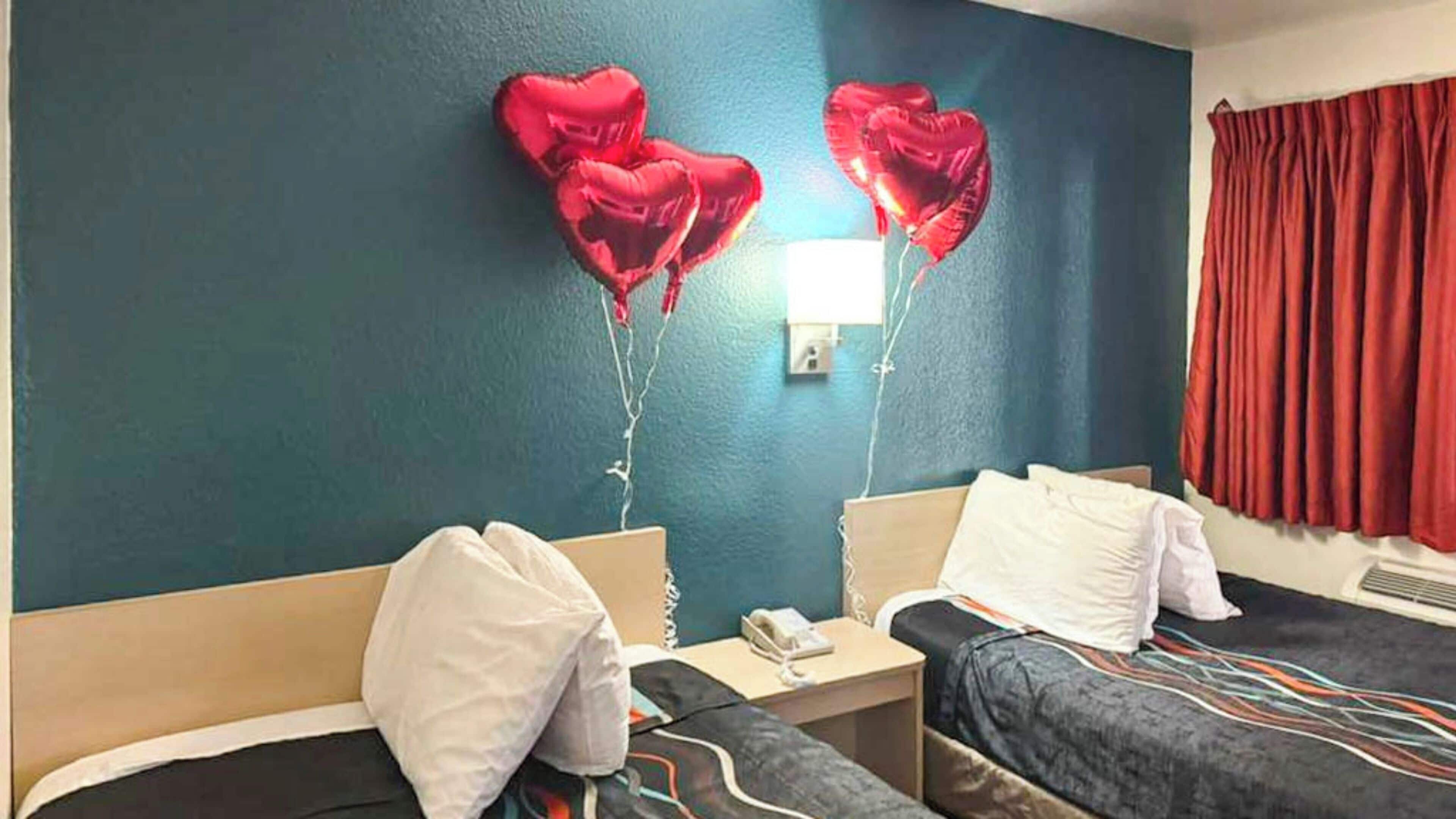Une chambre d’hôtel avec deux lits, une table de chevet avec un téléphone et des ballons en forme de cœur accrochés au mur.