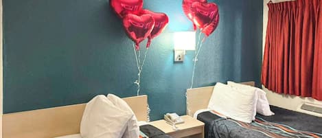 A hotel room with two beds, a nightstand with a phone, and heart-shaped balloons on the wall.