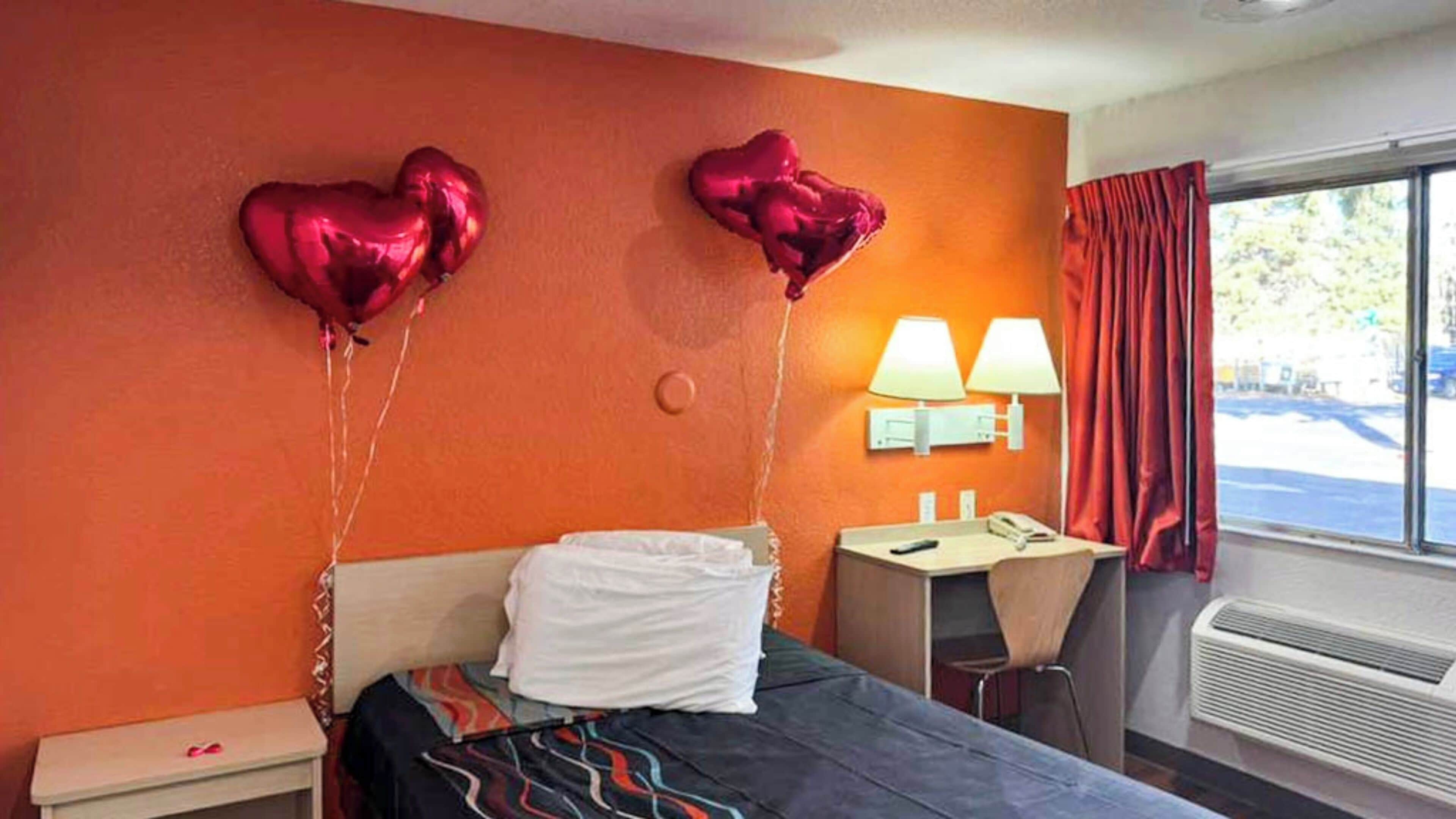 A hotel room with an orange wall, a bed with white bedding, a desk with a chair, two lamps, and a window with red curtains.