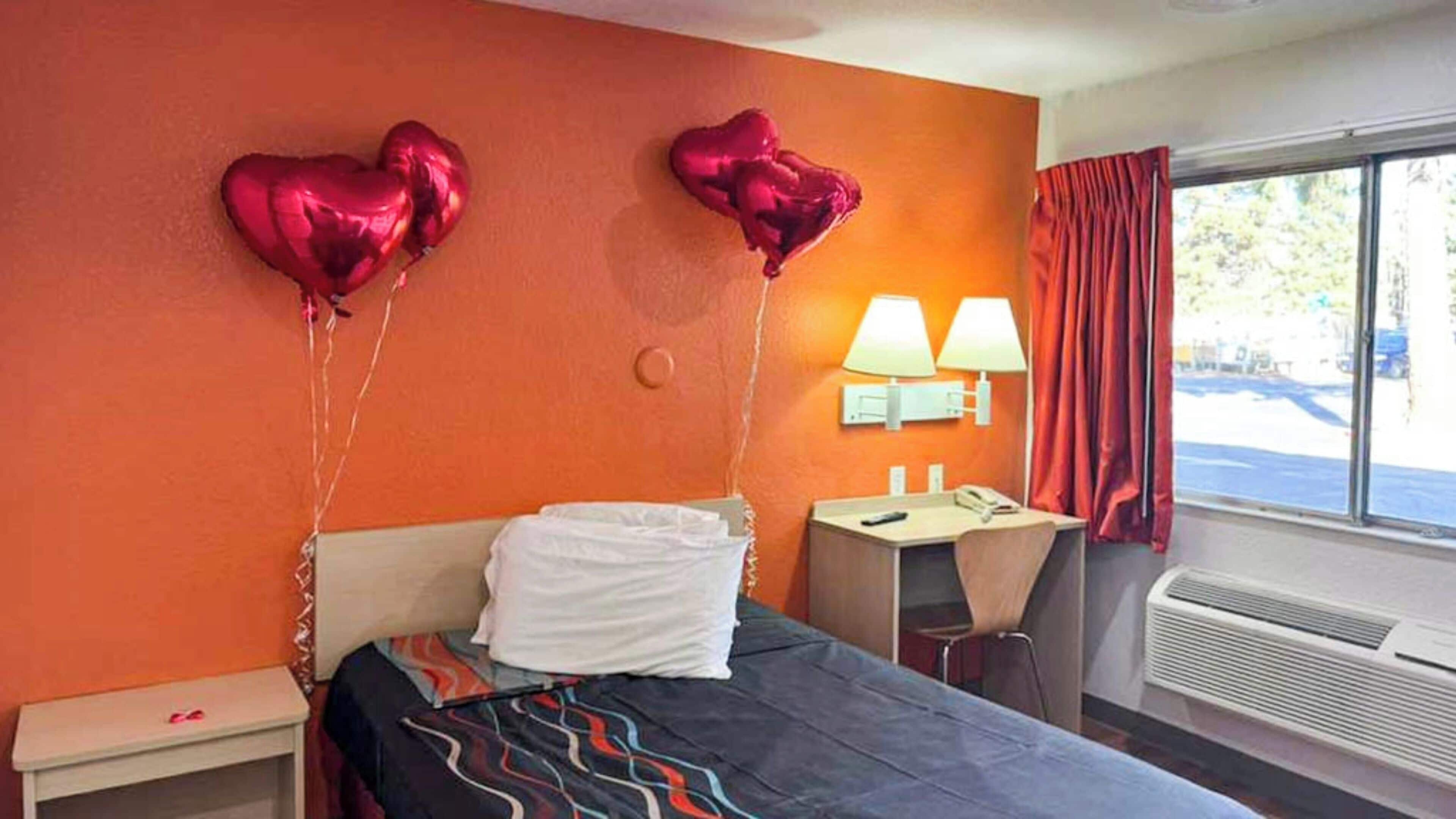 Habitación de hotel con pared naranja, cama con almohada blanca, escritorio con lámpara y dos globos con forma de corazón.