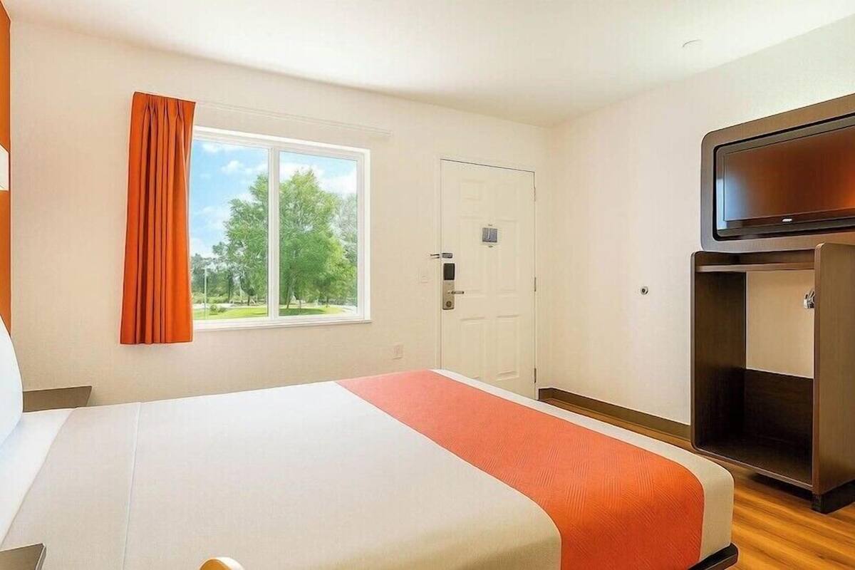 A hotel room with a bed, a television, a window with curtains, and a door.