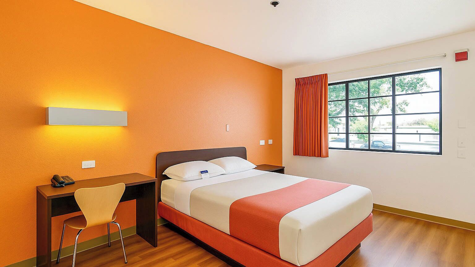 Una camera d'hotel con un letto, una scrivania con sedia, una finestra con tende e una lampada a parete.