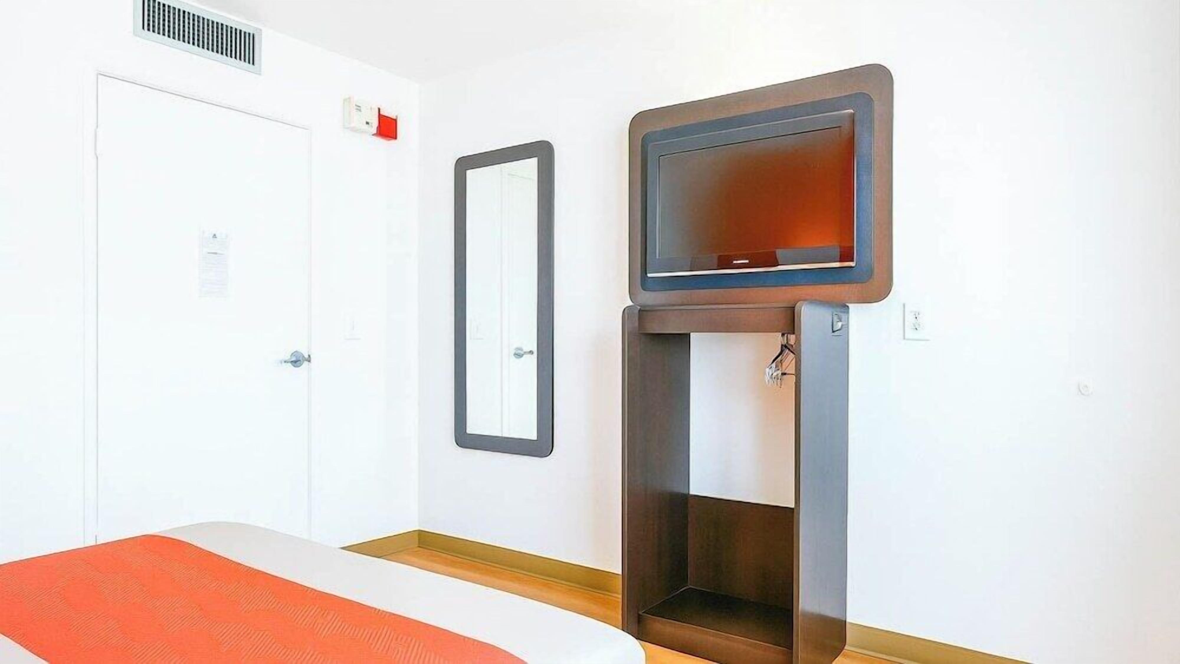A hotel room with a bed, a television mounted on a wooden stand, a white door, and a red carpet.