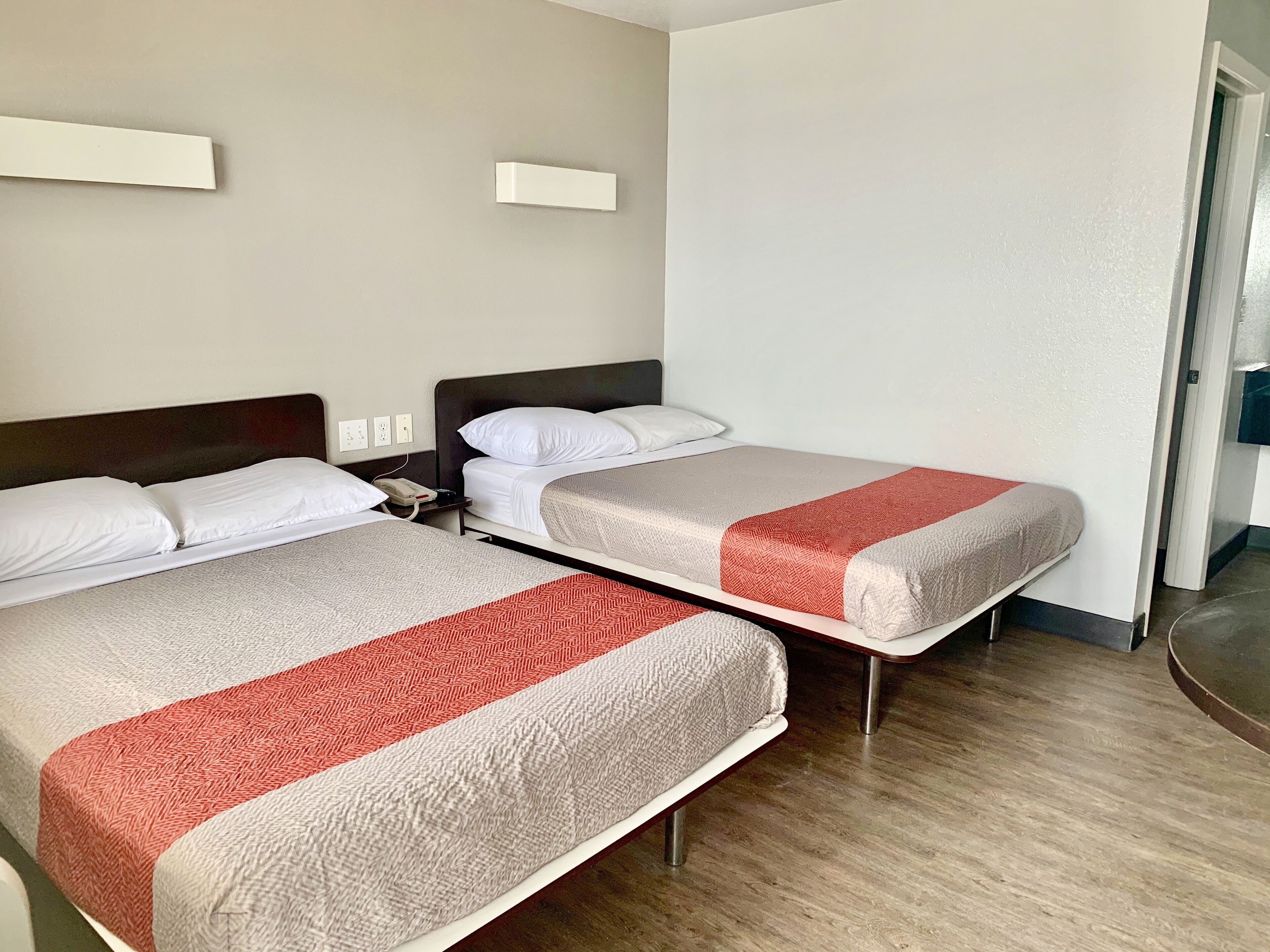 Two single beds with white and red bedding, a wooden floor, and a white wall with a small shelf.