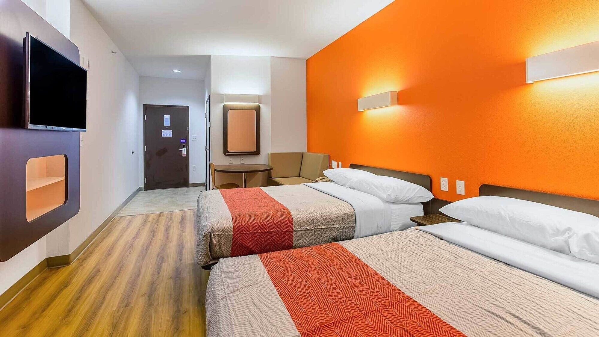 A hotel room with two beds, a TV, a desk, and an orange accent wall.