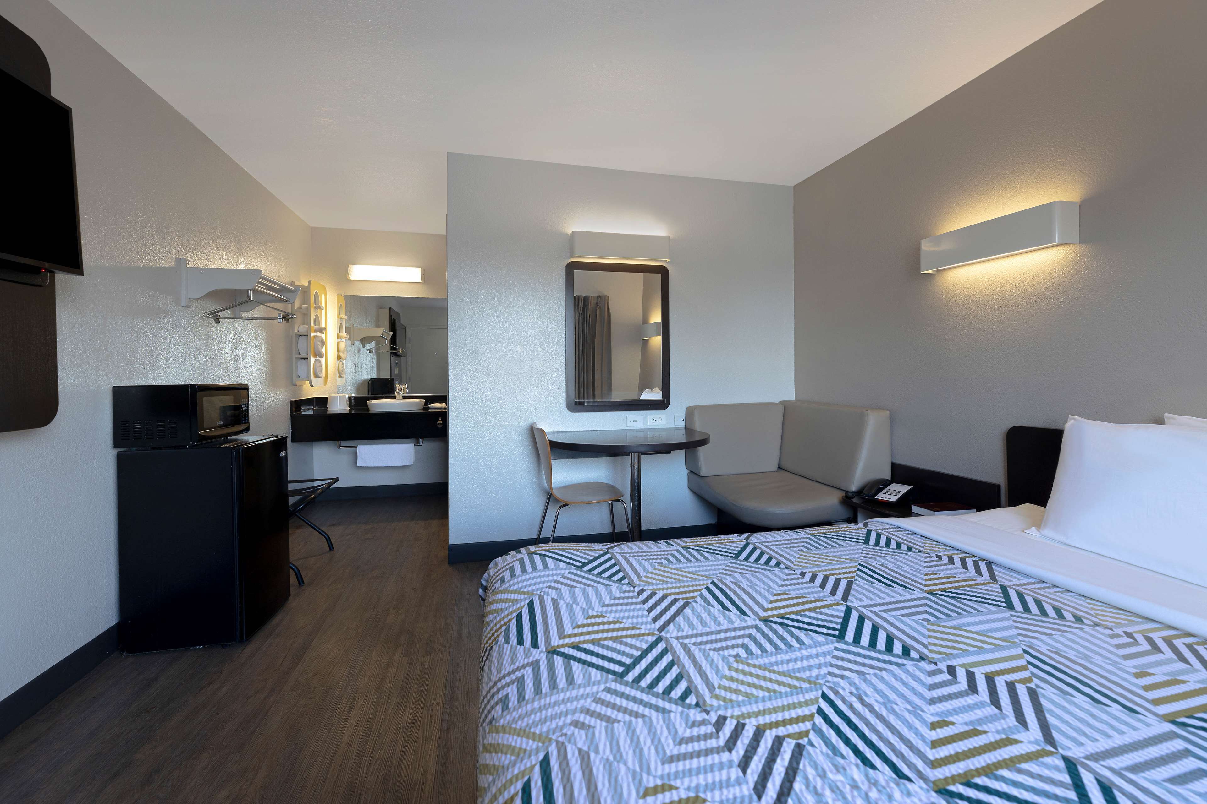 Habitación de hotel con cama, escritorio, silla, refrigerador pequeño, espejo y televisión montada en la pared.