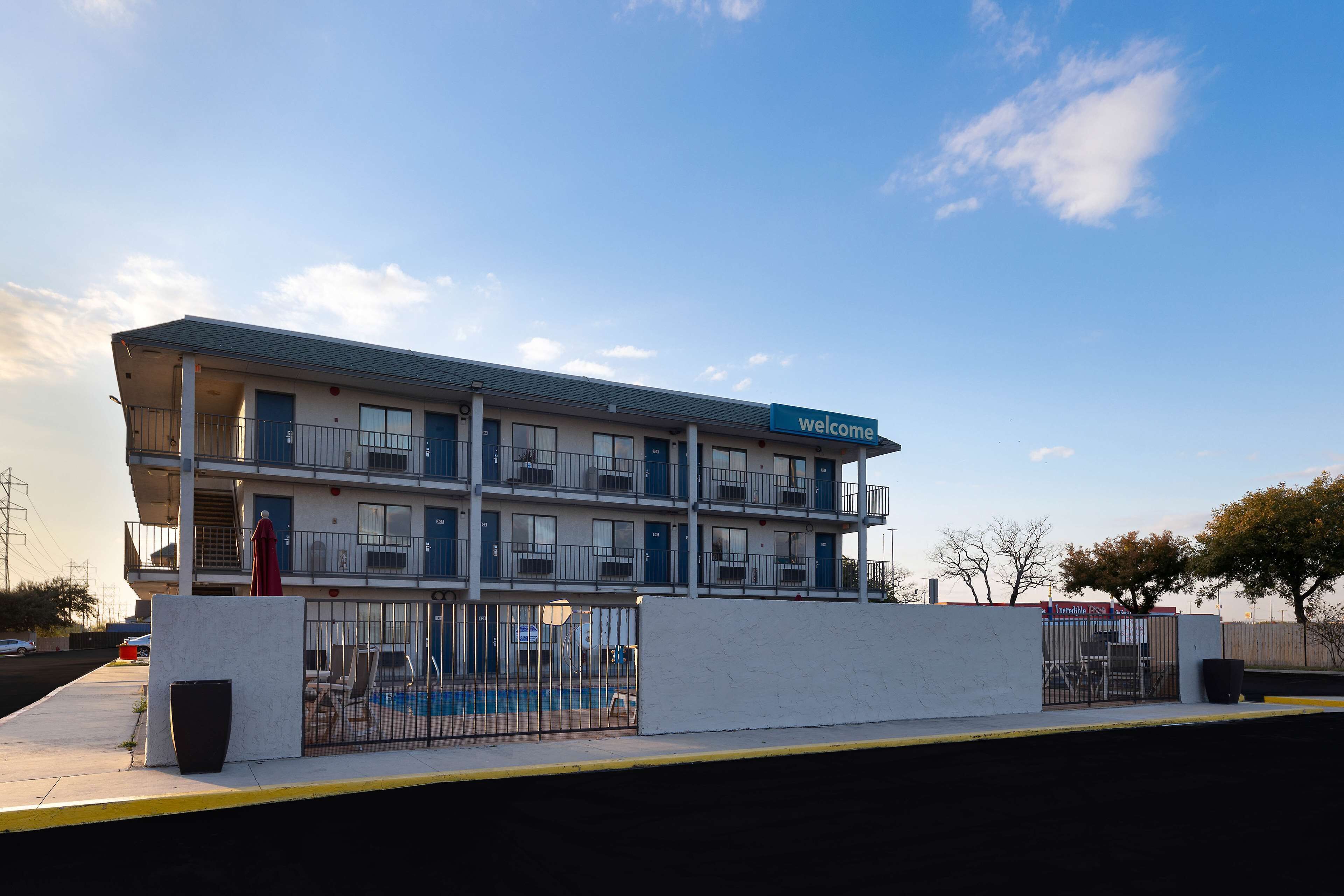 A multi-story motel with a 'Welcome' sign, a swimming pool, and a fenced parking area.
